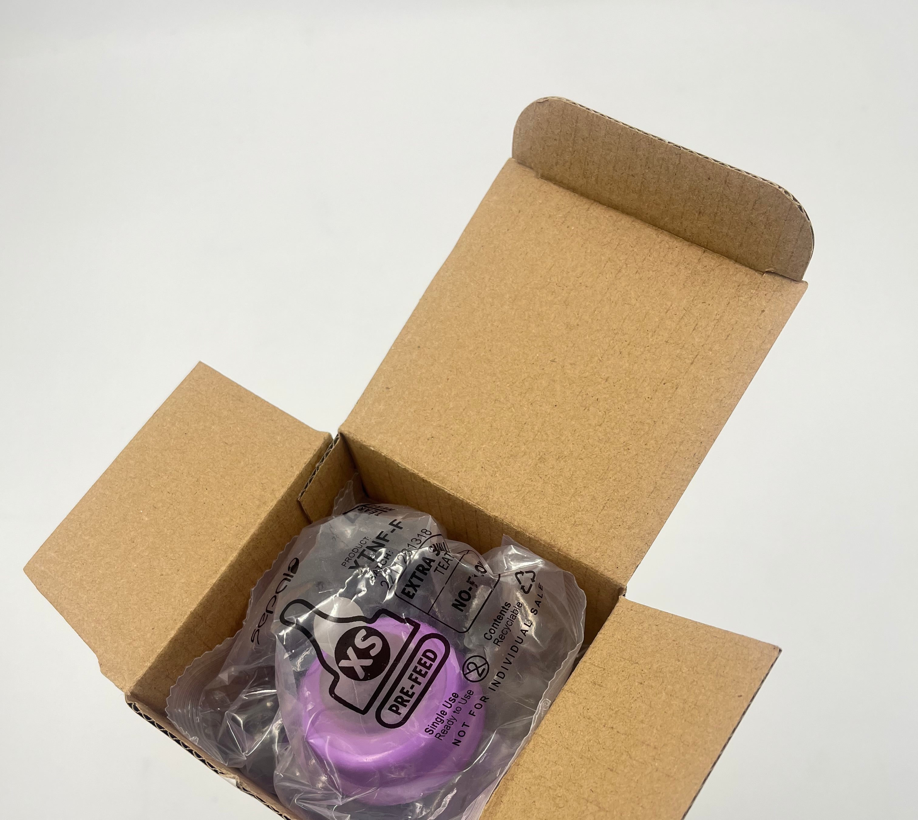 A packaged purple baby teat inside an open brown cardboard box on a plain white background.