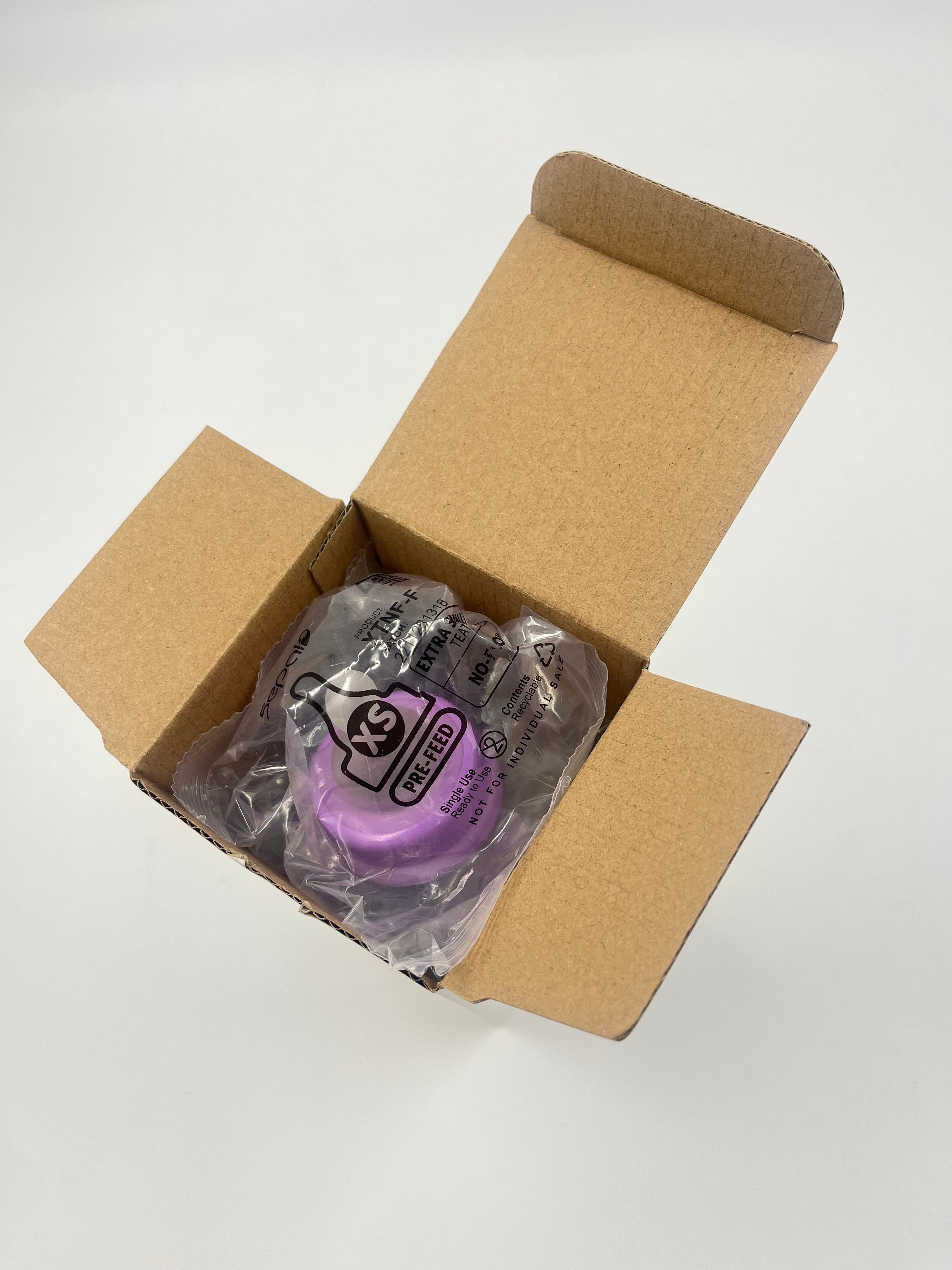A packaged purple baby teat inside an open brown cardboard box on a plain white background.